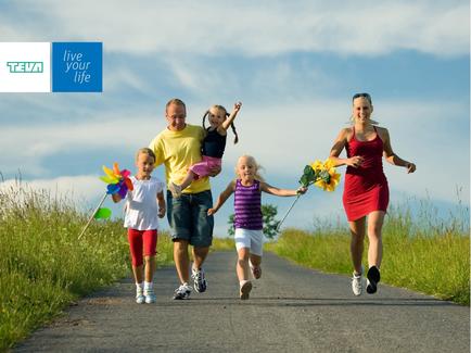 A family of five running on a road. The family consists of a father mother and two young children. The father is holding one of the children in his arms while the mother is running ahead of them. The children are holding sunflowers in their hands. The road is surrounded by tall grass and there is a blue sky in the background. On the top left corner of the image there is text that reads "Live your life".