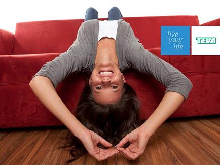 A young woman lying on her back on a red couch. She is upside down with her arms stretched out in front of her and her legs bent at the knees. She has long dark hair and is wearing a gray long-sleeved shirt and blue jeans. Her eyes are closed and she has a big smile on her face. The background is a wooden floor and there is a blue logo on the top right corner that reads "Live your life".