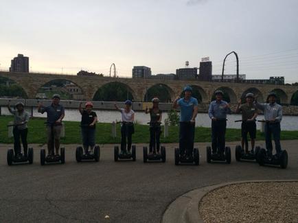 A group of nine people standing on seaways in a park. They are all wearing helmets and are posing for a photo. In the background there is a bridge with arches and a city skyline. The sky is blue and the sun is setting casting a warm glow over the scene. The people are smiling and appear to be enjoying themselves.