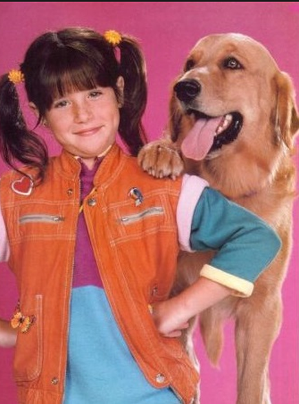 A portrait of a young girl and her golden retriever dog. The girl is wearing a blue dress and an orange vest with a heart-shaped patch on the left side. She has two yellow flower hair clips in her hair and is smiling at the camera. The dog is standing next to her with its tongue hanging out and its head tilted slightly to the side. The background is a solid pink color. The overall mood of the image is happy and playful.