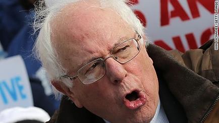 A close-up of Bernie Sanders an American politician speaking at a campaign event. He is wearing a brown jacket and glasses and appears to be in the middle of a speech. His mouth is open wide and his tongue is sticking out as if he is making a point. The background is blurred but it seems like he is surrounded by other people holding signs and banners. The image appears to have been taken at a political rally or event.