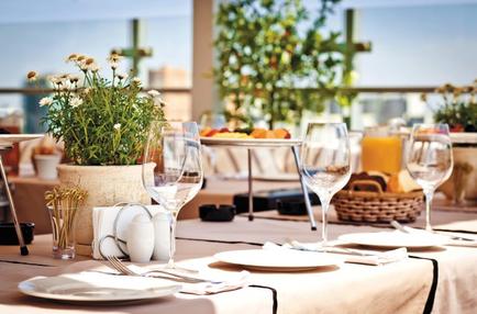 A table set up for a meal in a restaurant. The table is covered with a beige tablecloth and has white plates wine glasses and cutlery arranged neatly on it. There is a potted plant with white flowers on the left side of the table. On the right side there is a basket of fruit and a glass of orange juice. In the background there are other tables and chairs with plants and a view of the city skyline. The overall atmosphere of the restaurant is bright and airy.
