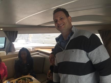A man standing in the middle of a boat smiling at the camera. He is wearing a grey and black striped sweater and a blue checkered shirt. Behind him there are two women sitting on a couch one of them is holding a plate of food and the other is looking at her phone. The boat appears to be on the water as there is a large window on the right side of the image. The interior of the boat is clean and well-lit.
