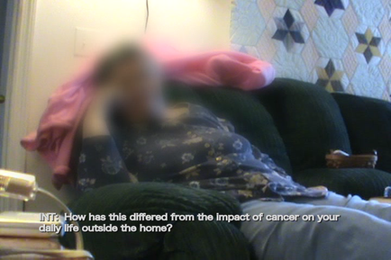 A person lying on a black couch with their head resting on a pink blanket. The person appears to be in a living room with a blue and white patterned wall in the background. The image is slightly blurred but it seems like the person is in motion. There is a text overlay on the image that reads "Int: How has this differed from the impact of cancer on your daily life outside the home?"