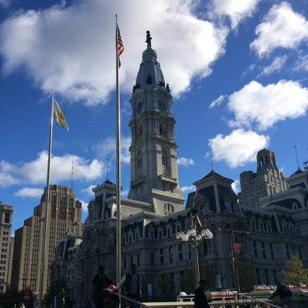 A large building with a clock tower in the center. The building appears to be a government building with multiple levels and ornate details. The clock tower has a dome on top and is decorated with statues and flags. The sky is blue with white clouds scattered across it. In front of the building there is a flagpole with the American flag flying in the wind. On the right side of the image there are other buildings and skyscrapers visible in the background. People can be seen walking on the sidewalk near the building.