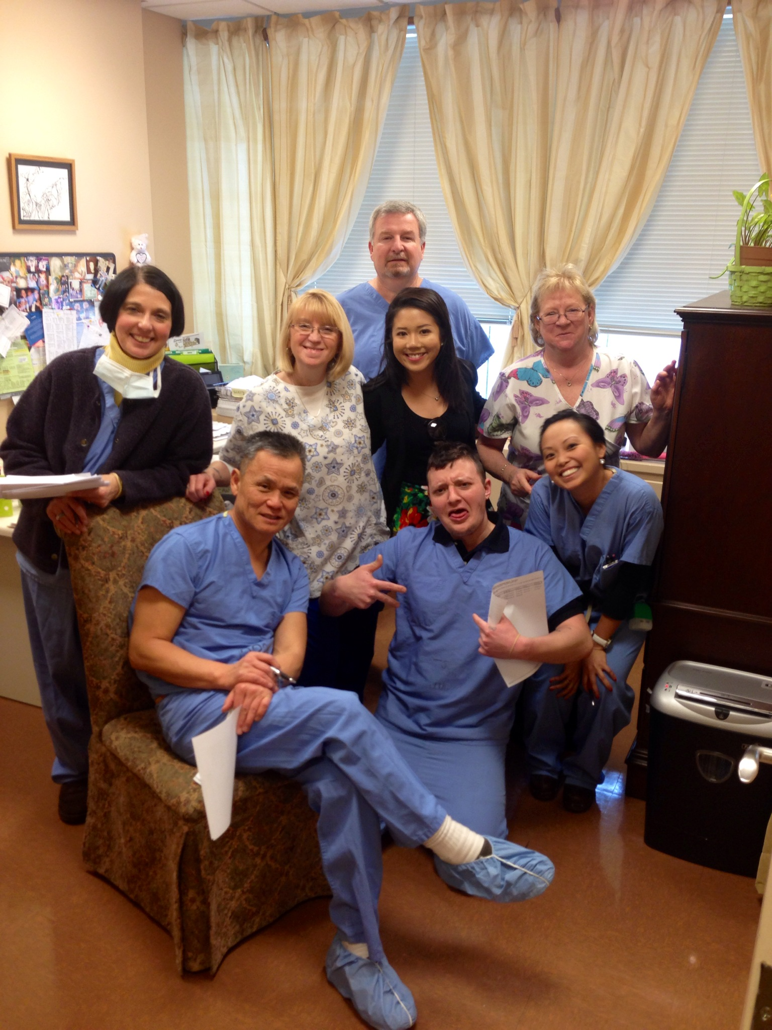A group of eight people posing for a photo in an office. Most are wearing blue scrubs and are standing in front of a window with white curtains. The man in the center is sitting on a chair with his legs crossed and is holding a piece of paper in his hand. The others are standing around him smiling and looking at the camera. On the left side of the image there is a woman with a face mask and holding a clipboard. In the background there is a bulletin board with pictures and papers pinned to it. The overall mood of the photo is happy and relaxed.