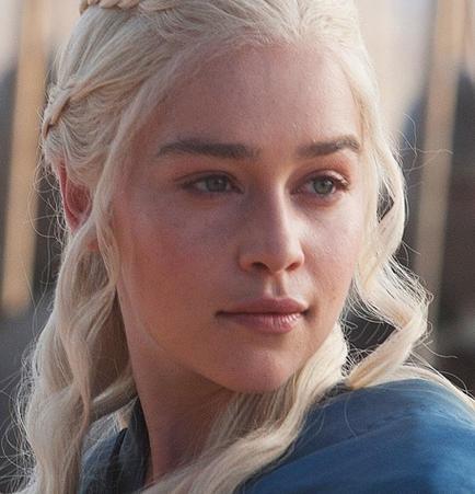 A close-up portrait of the character darner's Targaryen from the TV show Game of Thrones. She has long blonde hair styled in a braided updo and is wearing a blue dress. Her face is turned slightly to the side and she is looking directly at the camera with a serious expression. The background is blurred but it appears to be an outdoor setting with a wooden structure visible in the distance.