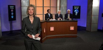 A news studio set with a woman standing in front of a desk. She is wearing a black suit and is holding a piece of paper in her hand. Behind her there are four men sitting at the desk all wearing suits and ties. The desk is made of wood and has a large screen on the right side with an image of a brain on it. The background is a purple wall with a large window. The woman appears to be a news anchor or a news reporter.