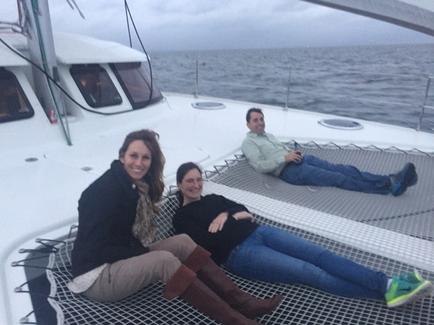 Three people sitting on the deck of a sailboat. The boat is white and the water is calm. The sky is overcast and the overall mood of the image is peaceful and serene. The person on the left is a young woman with long brown hair wearing a black jacket beige pants and brown boots. She is smiling and looking at the camera. Next to her is a man wearing a green jacket and blue jeans who is lying on his back with his legs crossed. He is also smiling and appears to be relaxed and enjoying the view. The woman in the middle is wearing a gray sweater blue jeans and green sneakers and the man on the right is wearing blue jeans. They are all wearing casual clothes and appear to be enjoying their time on the boat.