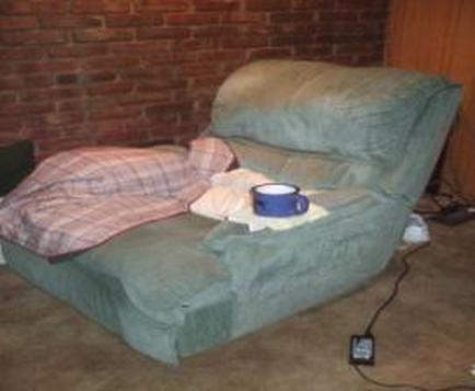 A green recliner chair in a room with a brick wall in the background. The chair is covered with a pink and white plaid blanket and there is a blue mug on the armrest. A white towel is resting on the chair next to it. A black power cord is plugged into a power outlet on the floor next to the chair.