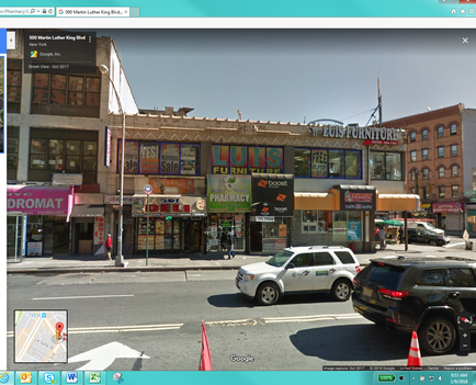 A street view of a busy intersection in a city. The intersection is located on a corner of a street with a large building on the left side of the image. The building has multiple shops and restaurants on the corner including a store called "lungs Furniture" and a Dromat. There are several other buildings on both sides of the street including one on the right side and another on the far left. The sky is blue and there are a few clouds in the sky. In the foreground there are two cars parked on the street and a traffic cone in front of the building. The image appears to be taken from a Google Street View window as there is a map visible in the bottom right corner.