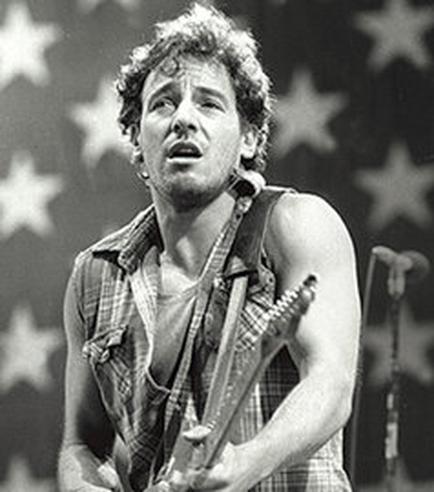 A black and white photograph of a young man playing an electric guitar on stage. He is wearing a plaid shirt and has curly hair. The background is blurred but it appears to be a stage with a large American flag. The man is looking up towards the sky with a focused expression on his face. There is a microphone on the right side of the image.
