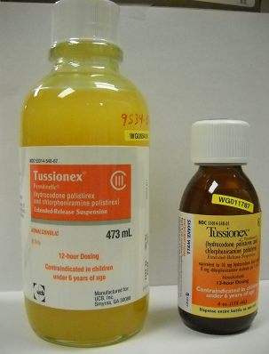 Two bottles of Tussionex. The bottle on the left is yellow in color and has a white label with the brand name Tussionex written in bold black letters. The label also has "473 ml" written on it in smaller black letters indicating that it is a 12-hour dosing and is contraindicated for children under 6 years of age. Next to the bottle there is a smaller bottle of Tussionex with a white cap and different branding. The smaller bottle has a yellow and orange label. Both bottles are placed on a white surface with a white background.