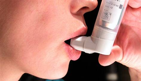 An extreme close-up of a person's face in profile with their mouth open as they use an inhaler device. The inhaler is both clear and white variously with a white label and black text. The (mostly black) background is blurred so the focus is on the person and the device.