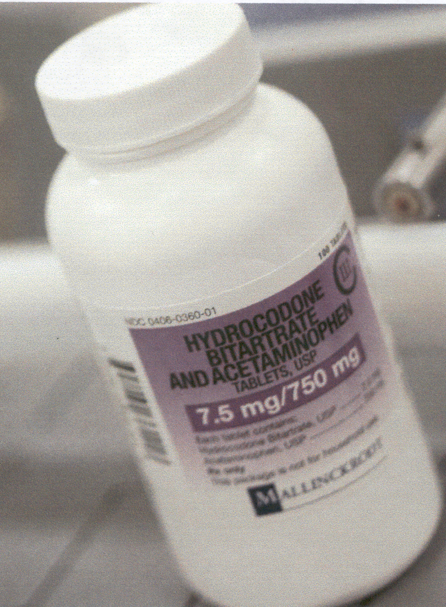 A close-up of a bottle of Hydrocodone Bitartrate and Acetaminophen tablets USP (7.5 mg/750 mg) CIII by Mallinckrodt. The bottle is white with a white and purple label with black and white text. The logo of Mallinckrodt is at the bottom of the label. The background (mostly gray) is blurred but appears to be a table.