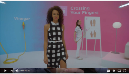 A screenshot from a video titled "Crossing Your Fingers". It shows a young woman with curly hair wearing a black and white checkered dress. She is standing in front of a pink background with the word "Vinegar" written in white. On the right side of the image there is a whiteboard with three illustrations of different types of fingers on it. The woman is holding a yellow skipping rope and appears to be in the middle of a dance move.
