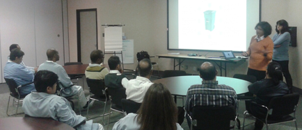 A group of people sitting around a round table in a conference room. They are all facing a projector screen and appear to be engaged in a presentation. The room has white walls and a door on the left side. On the right side of the image there is a woman standing in front of the screen giving a presentation to the group. She is wearing an orange sweater and is gesturing with her hands as she speaks. There is a laptop on the table and a whiteboard in the background. The people in the room are of different ages and genders and they are all looking towards the projector screen.