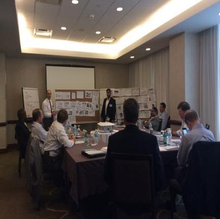 A group of people standing in a room with a whiteboard in the background. The room appears to be a conference room or a meeting room as there is a large window on the left side of the image. The people are wearing white shirts and are standing in front of the whiteboard which is covered with papers and notes. They are all facing the same direction and appear to be engaged in a discussion or brainstorming session. The floor is made of concrete and there are several circular lights on the ceiling creating a warm and inviting atmosphere.