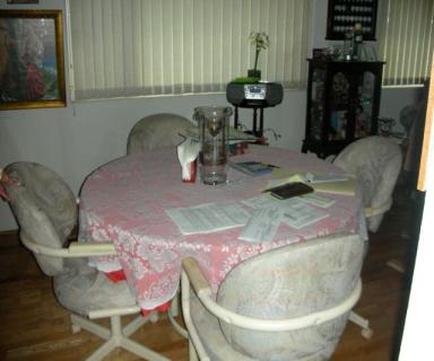 A dining room with a round table in the center. The table is covered with a pink tablecloth with a floral pattern. There are four chairs around the table each with a beige upholstered seat and backrest. On the table there are papers a glass of water and a few other items. In the background there is a window with white blinds and a painting hanging on the wall. To the right of the window there are shelves with various items on them. The room appears to be well-lit with natural light coming in from the window.