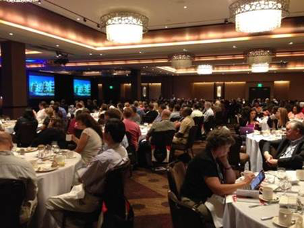 A large banquet hall with a high ceiling and chandeliers. The room is filled with people sitting at tables facing a large screen on the left side of the image. The tables are covered with white tablecloths and are set with plates cups saucers and glasses. The people are dressed in formal attire and appear to be engaged in a discussion or presentation. The walls are decorated with large windows and there is a door in the background. The overall atmosphere of the room is elegant and sophisticated.