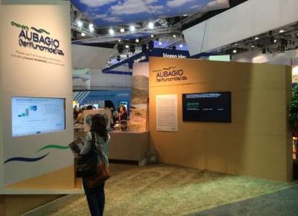 A trade show booth with a large screen on the left side of the booth. The booth is made of wood and has a beige color scheme. On the right side there is a large banner with the logo of adagio a company that provides information about the company. The banner also has a blue and white design with the company's name and logo. In the background there are other booths and people walking around the booth suggesting that the booth is part of a larger exhibition or trade show. A woman is standing in front of one of the booths looking at the screen.