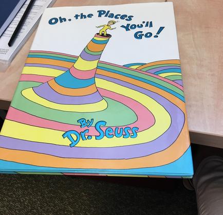 A photograph of a children book titled "Oh the Places You'll Go!" by Dr. Seuss. The book is resting on a wooden desk with a pen and a notebook next to it. The cover of the book features an illustration of a colorful striped cone with a yellow bird perched on top. The cone is surrounded by a rainbow-colored spiral pattern. The title is written in blue and yellow letters at the top of the cover.