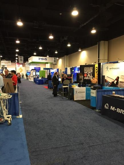 A large exhibition hall with a blue carpeted floor and white walls. There are several booths on the right side of the image each with a different design and color scheme. On the left side there are several people walking around and interacting with the booths. In the background there is a sign that reads "Insys" and a banner that says "Q-M-BR". The hall appears to be well-lit with bright lights hanging from the ceiling.