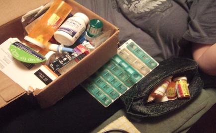 A cardboard box filled with various medical supplies. The box is open and the contents are spilling out onto the floor. On the left side of the box there are several bottles of different sizes and colors including orange white and green. Next to it there is a small black pouch with several small bottles of medicine inside. The pouch appears to be made of a dark material and has a zipper closure. The background is blurred but it seems like the person is sitting on a couch or a bed.