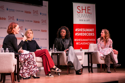 Four women sitting on a stage in front of a banner that reads "SHE Summit Rise & Lift". The women are engaged in a panel discussion with one woman holding a microphone and speaking to the audience. The woman on the left is wearing a black blazer and a floral skirt the woman in the middle is wearing red pants and a black top and the woman next to her is wearing black heels. The third woman is sitting in a beige armchair the fourth woman is in a pink dress and and the fifth woman is on the right. All four women appear to be of different ages and ethnicity's. The background is dark and there are water bottles on the stage.