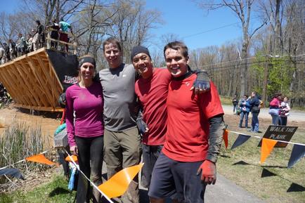 Four people two men and two women standing together and smiling at the camera. They are standing in front of a wooden structure that appears to be part of a race or obstacle course. The structure is made of wood and has a ramp on top. There are orange and black flags hanging from the sides of the structure. In the background there are trees and a crowd of people watching the race. The sky is blue and the ground is covered in grass.