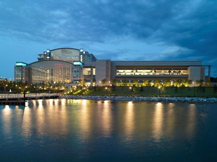 A night view of a large building with a curved glass facade. The building is located on the shore of a body of water with a dock on the right side. The sky is dark blue with a few clouds scattered across it. The water is calm and reflects the lights of the building and the surrounding buildings. There are trees and bushes along the shoreline and a few street lamps can be seen on the buildings. The overall atmosphere of the image is peaceful and serene.