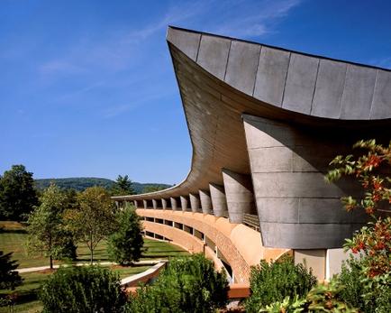 A modern building with a unique architectural design. The building is made of concrete and has a curved roof that extends from the top to the bottom. The roof is made up of multiple layers of concrete panels that create a wave-like pattern. The panels are arranged in a way that creates a sense of depth and dimension. The building is surrounded by a well-manicured garden with trees and shrubs. The garden is well-maintained with a variety of colorful flowers and plants. In the background there are rolling hills and a clear blue sky. The overall atmosphere of the image is peaceful and serene.