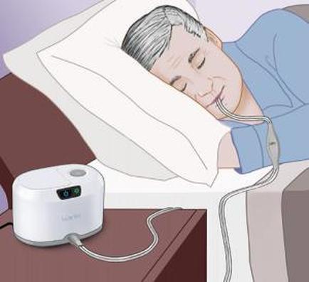A man sleeping in bed with his eyes closed and his head resting on a white pillow. He is wearing a blue shirt and appears to be in a deep sleep. On the right side of the image there is a white device with a digital display and a cable connected to it. The device is likely a sleep apnea machine which is used to help reduce the amount of sleep in the man's body. The background is a plain purple wall.