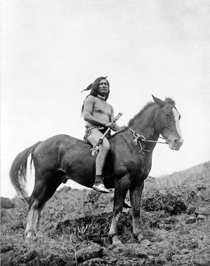 A black and white photograph of a Native American man sitting on top of a horse. The man is wearing a traditional headdress and is holding a spear in his right hand. He is sitting on the back of the horse which is standing on a rocky hillside. The horse is a dark brown color with a white mane and tail. The background is a barren landscape with patches of grass and shrubs. The sky is overcast and the overall mood of the image is somber.