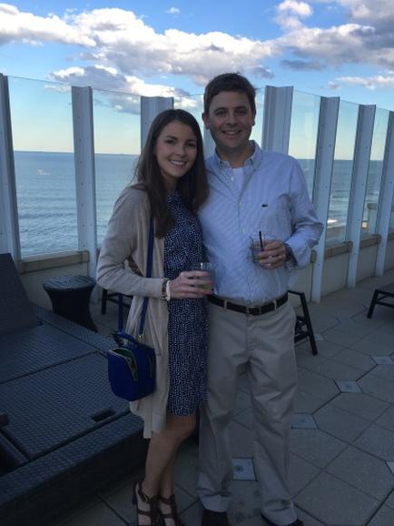 A young couple standing on a balcony overlooking the ocean. They are both smiling and holding drinks in their hands. The woman is wearing a blue dress with white polka dots and a beige cardigan while the man has a white shirt and khaki pants. The balcony has a glass railing and there are lounge chairs and tables scattered around. The ocean can be seen in the background with a clear blue sky above.