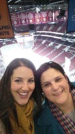 A selfish of two young women standing in front of a shopping mall. They are both smiling and looking directly at the camera. The woman on the left is wearing a yellow scarf and a blue jacket while the woman in the middle has long dark hair. The background shows a shopping cart with various items on it including a sign that reads "Alpha Outlets". The mall appears to be bustling with activity with people walking around and cars parked in the background.