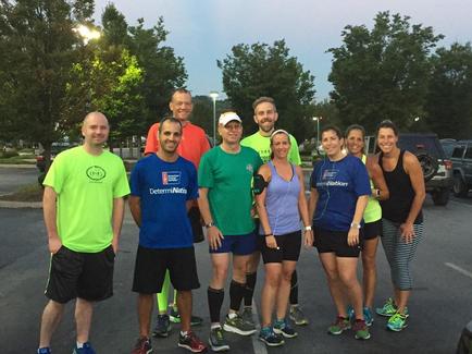 A group of nine people standing in a parking lot. They are all smiling and appear to be happy and relaxed. The group is composed of men and women of different ages and ethnicity's. Some of them are wearing casual clothes while others are wearing t-shirts with logos or designs. The parking lot is surrounded by trees and there are cars parked in the background. The sky is blue and the sun is setting casting a warm glow over the scene.