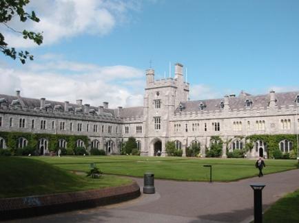 A large grand building with a castle-like appearance. The building is made of stone and has multiple levels with arched windows and turrets. The exterior of the building is covered in ivy and there is a well-manicured lawn in front of it. The sky is blue and there are a few clouds scattered across it. In the foreground there are two people walking on the path leading up to the building. The overall atmosphere of the image is peaceful and serene.
