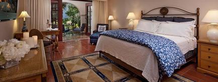 A luxurious bedroom with a large king-sized bed in the center. The bed has a black metal headboard and is covered with a blue and white patterned comforter and pillows. There are two nightstands on either side of the bed with lamps on them. On the left side there is a wooden dresser with a vase of white flowers and a flat-screen TV mounted on the wall. The room has a tiled floor and a large window with white curtains. In the background there are two armchairs and a balcony with a view of the ocean. The overall style of the room is elegant and inviting.