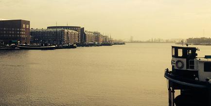 A photograph of a harbor with several boats and buildings on the water. The sky is overcast and the water is calm. On the right side of the image there is a tugboat docked at a pier. The tugboat is white with a black roof and has a small window on the front. In the background there are several buildings and a bridge. The image is taken from a low angle looking down on the harbor.