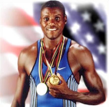A young African-American man holding two gold medals in his hands. He is wearing a blue and white striped tank top and has a big smile on his face. The background is blurred but it appears to be an American flag. The man looks proud and happy likely after winning a gold medal at a sporting event.