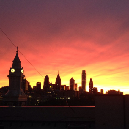 A photograph of a city skyline at sunset. The sky is a beautiful orange and pink color with the sun setting in the background. The buildings in the skyline are silhouetted against the sky creating a stunning contrast of colors. In the foreground there is a tall clock tower with a steeple and a cross on top. The clock tower is lit up casting a warm glow over the city. The city appears to be New York City as there are several skyscrapers visible in the distance. The overall mood of the image is peaceful and serene.