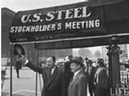 A black and white photograph of a group of people standing in front of a sign that reads "U.S. Steel Stockholder's Meeting." The sign is rectangular in shape and has a large bold font. The people in the photograph are dressed in formal attire with some wearing suits and others wearing hats. One person is holding a microphone and appears to be giving a speech or presentation. The background shows a building and a fence suggesting that the photo was taken at a public event or gathering.