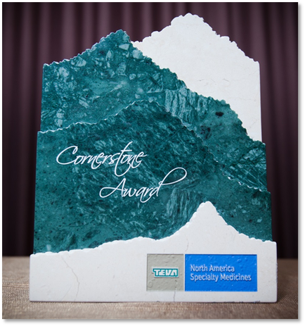 A photograph of a trophy or award for a company named "Cornerstone Award". The trophy is made of a white base with a blue and green marble-like texture. The top half of the trophy is shaped like a mountain peak with the words "North America Specialty Medicines" written in a cursive font in the center. The bottom half is a darker shade of blue with a small logo of the company's name in the bottom right corner. The background is a maroon curtain.