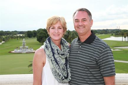 A man and a woman standing on a balcony overlooking a golf course. They are both smiling and posing for the camera. The woman is on the left side of the image wearing a white tank top and a blue scarf with a floral pattern. She has blonde hair and is looking directly at the camera with a slight smile on her face. The man on the right side is wearing a striped polo shirt and is also smiling. In the background there is a beautiful view of the golf course with a fountain and trees. The sky is blue and there are a few clouds in the distance.
