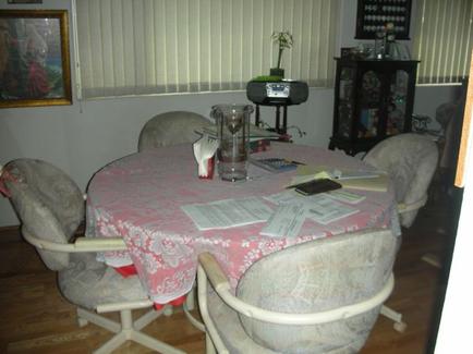 A dining room with a round table in the center. The table is covered with a pink tablecloth with a floral pattern. There are four chairs around the table each with a beige upholstered seat and backrest. On the table there are papers a glass of water a calculator and a few other items. In the background there is a window with white blinds and a painting hanging on the wall. To the right of the window there are shelves with various decorative items. The floor is made of hardwood.
