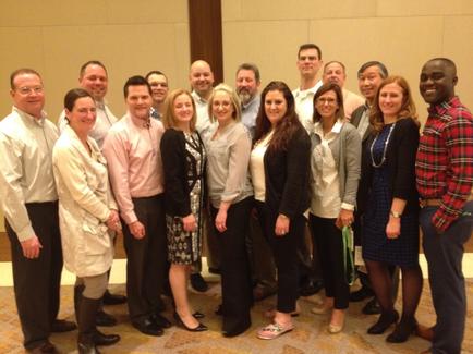 A group of people standing in a room with a beige carpeted floor and a wooden wall in the background. There are around 20 people in the group all of them are smiling and looking at the camera. They appear to be of different ages and ethnicity's. Some are wearing casual clothes while others are dressed in formal attire. The group is arranged in a semi-circle with some people standing on the left side of the image and others on the right side.