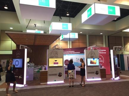 A trade show booth with three people standing in front of it. The booth is decorated with white and green banners with the letters "CNS" on them. There are three large screens on the booth each displaying a different image and text. The people in the image are wearing casual clothing and appear to be engaged in conversation. In the background there are other booths and people walking around the booth. The overall atmosphere of the booth is modern and professional.