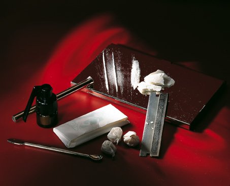 A set of tools on a red background. On the left side of the image there is a black spray bottle with a long handle and a nozzle. Next to the spray bottle there are two small white objects one of which appears to be a piece of paper or a napkin and the other of which is a metal tool with a curved handle. The metal tool is resting on top of a red surface which is covered in a layer of white powder. There are also a few baggies of an unidentified white powder scattered around the tools. The overall color scheme of the background is red and black creating a striking contrast with the white objects.