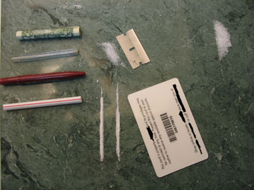 A green countertop with various items scattered on it. On the left side of the countertop there are two red and white pens one with a green cap and the other with a white cap. Next to the pens there is a small piece of paper with a barcode and some text on it which appears to be a label or a bar code. In the center of the image there appears to have a small amount of white powder scattered around. The items are arranged in a scattered manner with some of them overlapping each other.